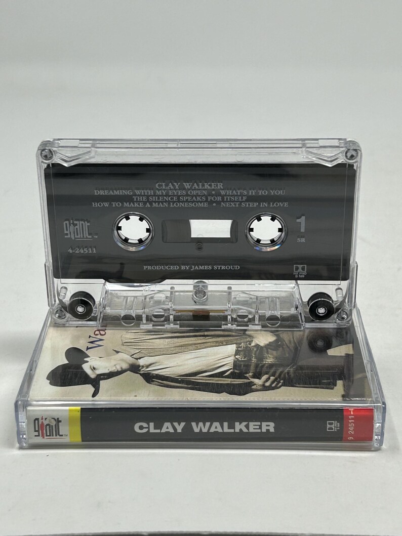 May include: A clear cassette tape featuring Clay Walker's album. The tape has a black label with white text listing song titles like "Dreaming With My Eyes Open." The cassette case is open, revealing the tape and album art. The album art features a black and white image of a man.