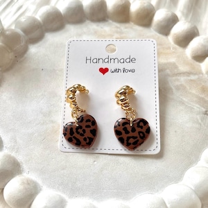 Puede incluir: Pendientes en forma de corazón con estampado de leopardo. Los pendientes son marrones con manchas negras y detalles metálicos dorados. La tarjeta detrás de los pendientes dice "Handmade with Love."