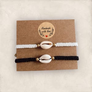 Può includere: Due braccialetti fatti a mano con cordoncino bianco e nero, ognuno con un ciondolo a conchiglia bianca e perline dorate. I braccialetti sono esposti su una carta marrone con un adesivo che dice "Fatto a mano con amore".