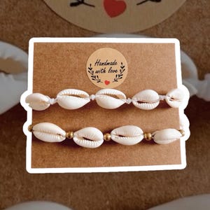 Può includere: Due braccialetti di conchiglie bianche su una carta marrone con un adesivo che dice "Fatto a mano con amore".