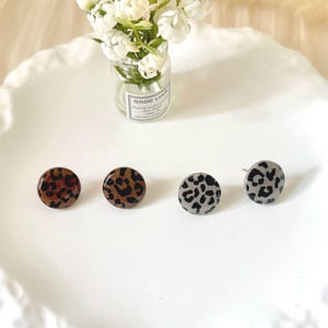 Può includere: Quattro orecchini a bottone rotondi con motivi leopardati marroni, neri e grigi. Gli orecchini sono esposti su un piatto bianco smerlato, con un piccolo vaso di fiori bianchi sullo sfondo.