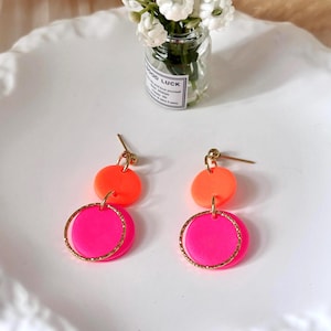 Könnte beinhalten: Ein Paar neonpinke und orangefarbene Ohrringe. Jeder Ohrring hat eine kleine orangefarbene Scheibe, eine größere pinkfarbene Scheibe und einen goldfarbenen Reifen. Die Ohrringe werden auf einer weißen, strukturierten Oberfläche präsentiert.