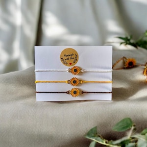 Könnte beinhalten: Drei Freundschaftsarmbänder mit Sonnenblumenanhängern. Die Armbänder sind weiß, gelb und braun. Die Karte lautet "Handgefertigt mit Liebe".