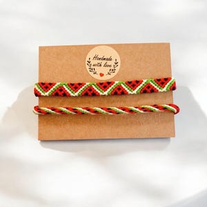 Könnte beinhalten: Zwei handgefertigte Freundschaftsarmbänder mit einem Wassermelonenmuster in Rot, Grün und Schwarz. Die Armbänder befinden sich auf einer braunen Karte mit einem Aufkleber, auf dem "Handgefertigt mit Liebe" steht.
