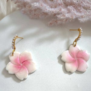 Könnte beinhalten: Ein Paar Ohrringe mit Blumenmotiv. Jeder Ohrring hat einen weißen und rosa Blütenanhänger, einen goldfarbenen Stecker und einen geschwungenen Akzent, der mit kleinen, weißen Perlen verziert ist. Die Ohrringe werden auf einer weißen Oberfläche präsentiert.