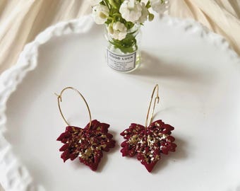 May include: Burgundy leaf earrings with gold detailing, suspended from gold hoops. The earrings are displayed on a white plate. A small glass jar with white flowers and the words "GOOD LUCK" is in the background.