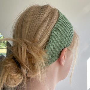 Op de afbeelding: Een groene gebreide hoofdband met een geribbelde textuur. De hoofdband wordt door een persoon gedragen.