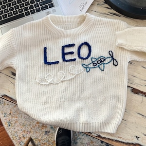 Puede incluir: Suéter de punto blanco con letras bordadas en azul que dicen "LEO" y un avión azul con una hélice blanca. El avión está bordado con una línea blanca que se arrastra detrás de él.