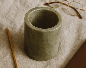 May include: An olive-green stone pencil holder, cylindrical in shape, rests on a neutral linen surface. A wooden pencil and gold-rimmed spectacles are also present. A brown notebook is partially visible, suggesting a workspace.