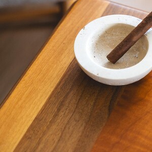 May include: A brown cigar rests in a small, off-white ashtray. The ashtray is circular and sits on a wooden table with a warm, reddish-brown finish. The scene suggests a moment of relaxation.