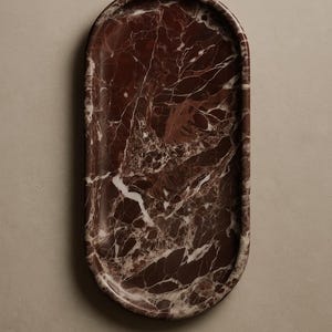 Puede incluir: Una bandeja ovalada de m&aacute;rmol marr&oacute;n. La bandeja tiene un acabado pulido y un patr&oacute;n de m&aacute;rmol natural con vetas blancas. Un art&iacute;culo decorativo para el hogar.