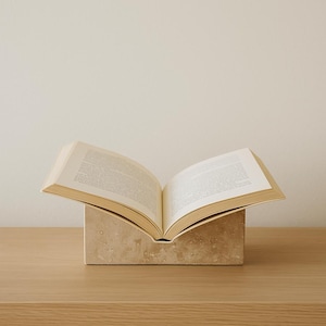 May include: An open book rests on a beige stone book stand. The book's pages are visible, displaying printed text. The stand is rectangular with a curved top, placed on a light wood surface. The background is a plain, off-white wall.