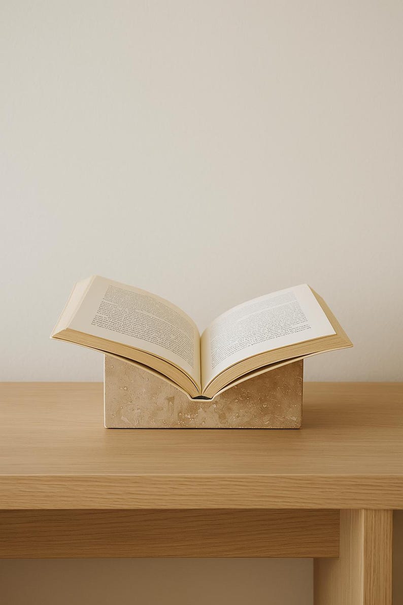 May include: An open book rests on a beige stone book stand. The book's pages are visible, displaying printed text. The stand is rectangular with a curved top, placed on a light wood surface. The background is a plain, off-white wall.