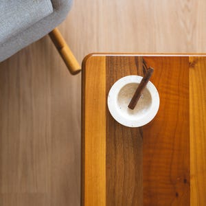 May include: A top-down view of a wooden table with a small white bowl and a cinnamon stick. The table has a natural wood grain pattern with varying shades of brown and orange. A gray armchair is partially visible in the background.