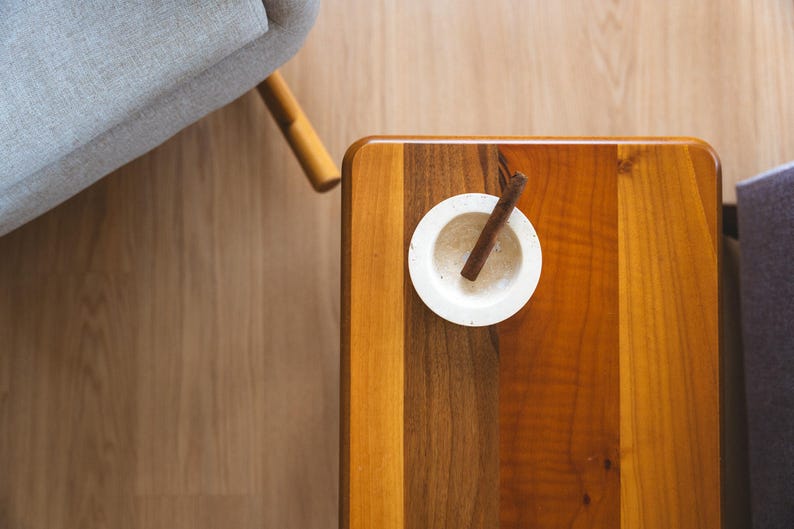 May include: A top-down view of a wooden table with a small white bowl and a cinnamon stick. The table has a natural wood grain pattern with varying shades of brown and orange. A gray armchair is partially visible in the background.