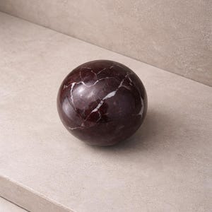 May include: A polished, spherical decorative object. The sphere is a deep burgundy color with white veining throughout. The marble ball is resting on a light beige surface. This decorative item could be used as a paperweight or home decor.