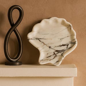 May include: A decorative marble seashell-shaped tray with dark veining sits on a white surface. A dark bronze sculpture in the shape of an infinity symbol is to the left of the tray. The background is a solid tan color.