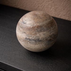 May include: A spherical decorative stone object with a marbled appearance. The stone features a mix of brown, tan, and gray tones, creating a natural, earthy aesthetic. The sphere is resting on a dark surface.
