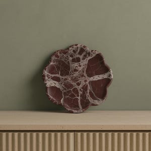 Può includere: Un vassoio decorativo a forma di fiore, in marmo bordeaux con venature bianche. Il vassoio è appoggiato su un mobile chiaro con dettagli a coste verticali, contro un muro verde tenue.