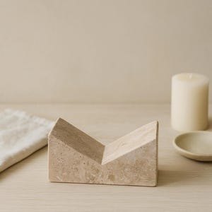 May include: A beige, geometric stone object with a V-shaped design. The object is on a light wood surface, with a candle and small dish in the background. The stone object has a textured surface.
