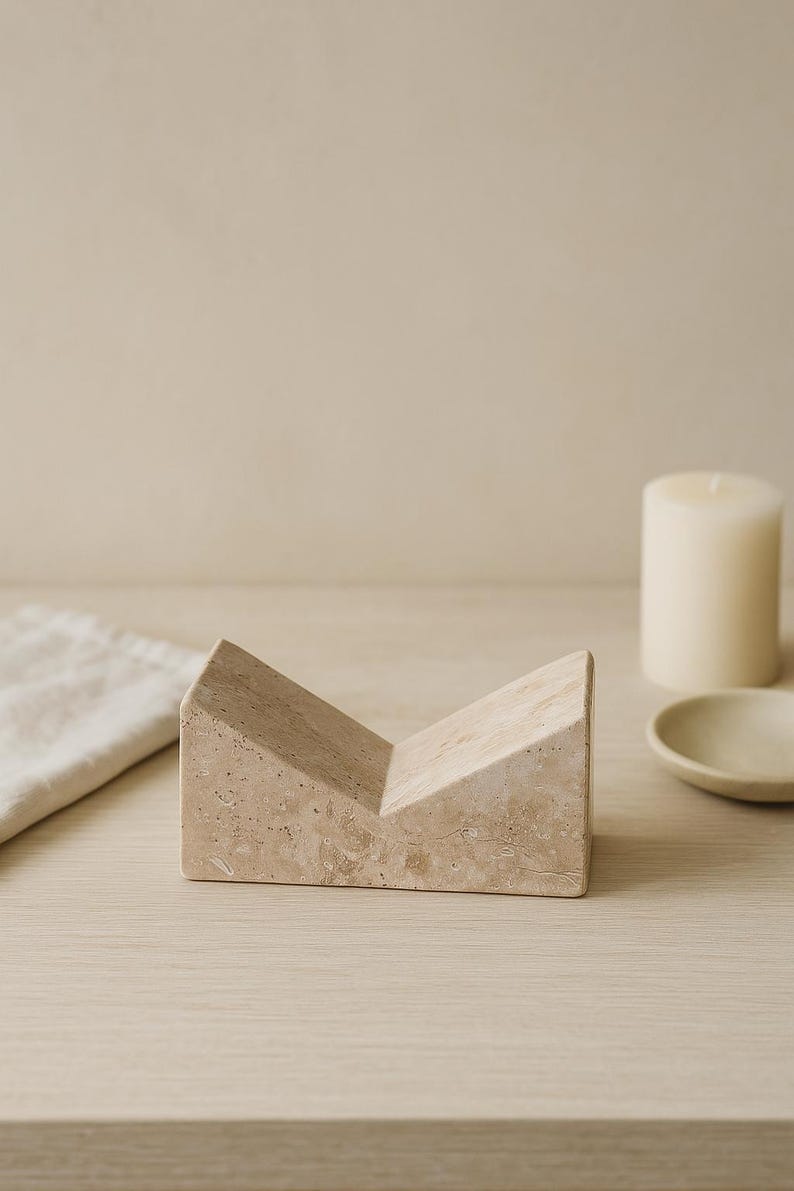 May include: A beige, geometric stone object with a V-shaped design. The object is on a light wood surface, with a candle and small dish in the background. The stone object has a textured surface.
