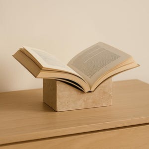 May include: An open book resting on a beige, square book stand. The pages of the book are visible, with text printed on them. The book and stand are on a light-colored wooden surface.