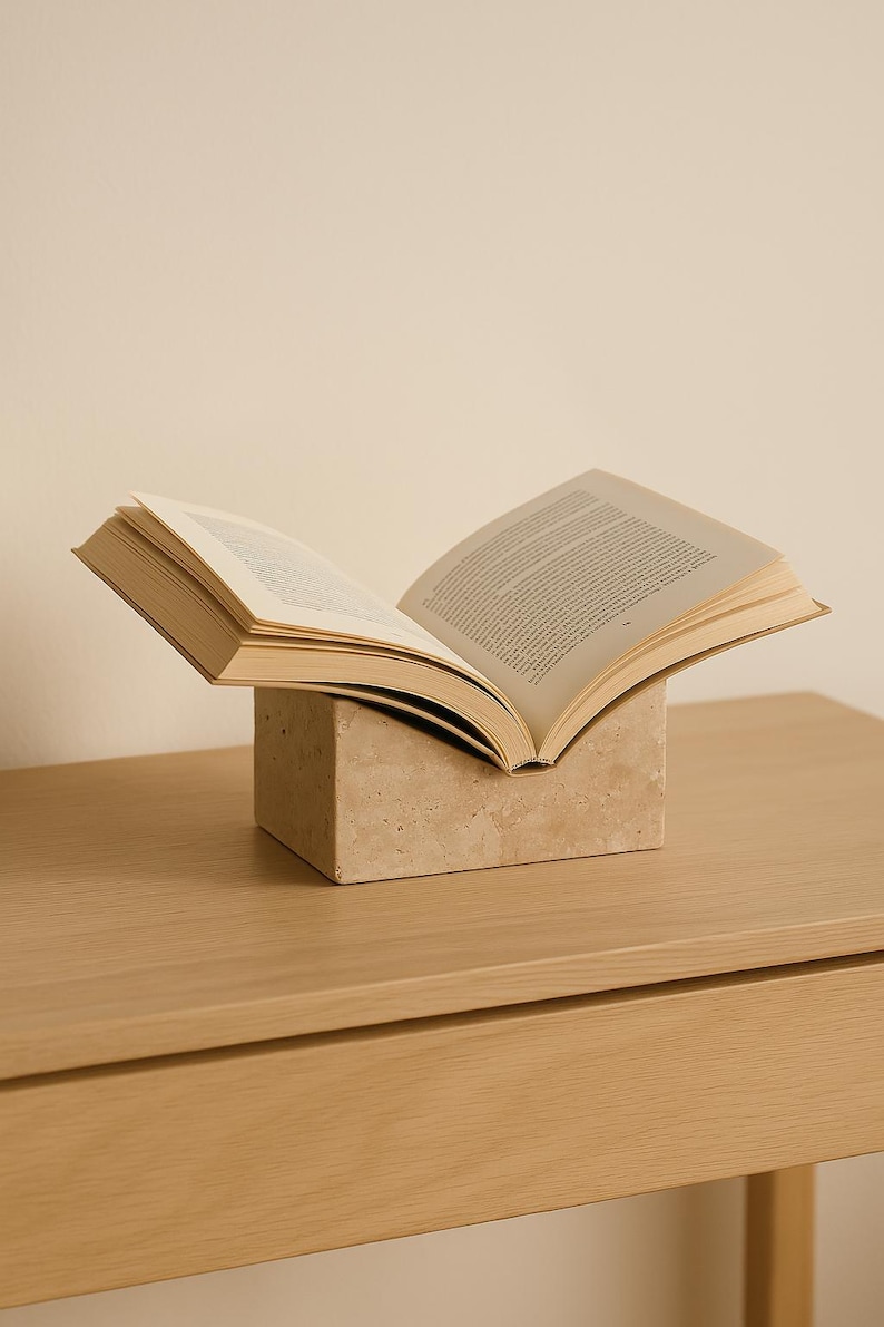 May include: An open book resting on a beige, square book stand. The pages of the book are visible, with text printed on them. The book and stand are on a light-colored wooden surface.