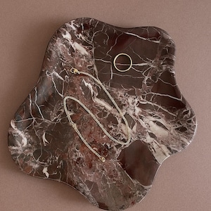 Könnte beinhalten: Dekorative Schale mit organischer Form aus dunkelrotem Marmor mit weißen Adern. Eine goldene Halskette und ein goldener Ring liegen auf der Schale und bilden eine minimalistische Schmuckpräsentation.