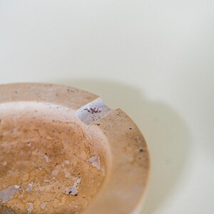 May include: A close-up of a round, beige ashtray with a textured surface. The ashtray has a small notch for resting cigarettes. The ashtray is made of a stone-like material and has a rustic appearance.