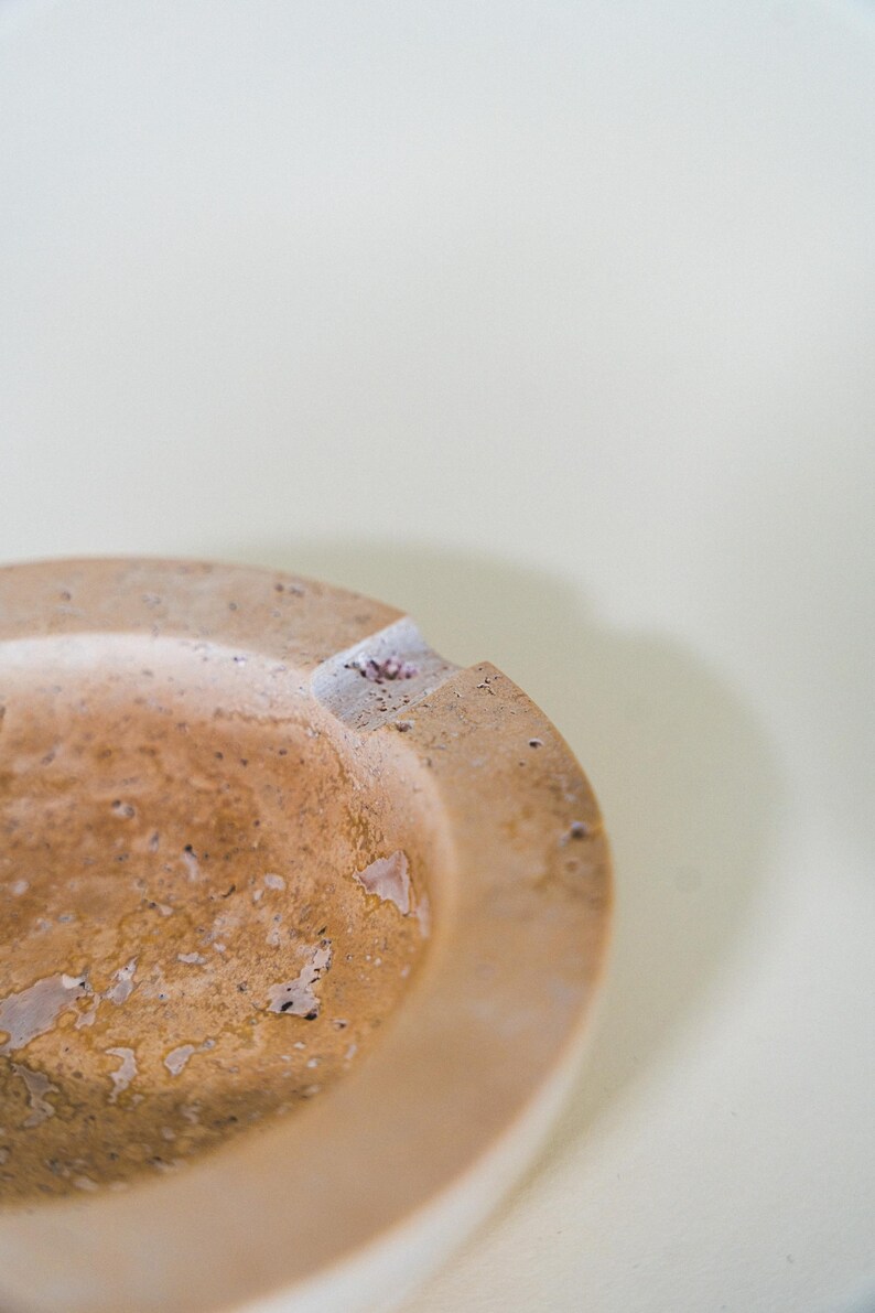 May include: A close-up of a round, beige ashtray with a textured surface. The ashtray has a small notch for resting cigarettes. The ashtray is made of a stone-like material and has a rustic appearance.