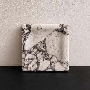 Puede incluir: Bandeja cuadrada de mármol con base blanca y vetas moradas oscuras. La bandeja está colocada sobre una superficie negra con una pared blanca texturizada en el fondo. Esta bandeja decorativa es ideal para servir o exhibir objetos pequeños.