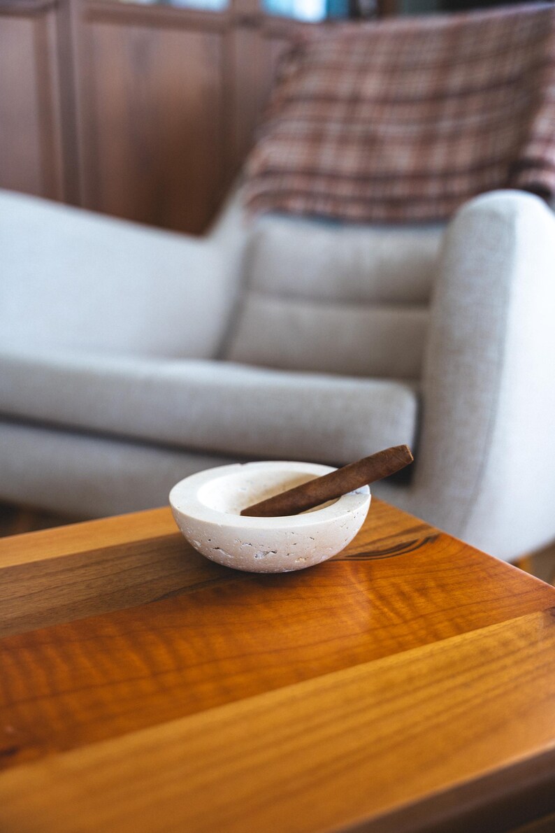May include: A white, textured ashtray with a cigar resting inside, placed on a wooden table. The ashtray is round with a shallow bowl. The cigar is brown. The table has a warm, reddish-brown finish. A light-colored armchair is in the background.
