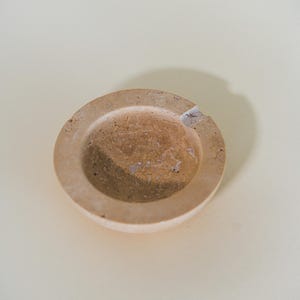 May include: A round, beige ashtray with a shallow bowl and a raised rim. The ashtray has a natural, textured appearance with a small notch for resting cigarettes. The ashtray is made of a light-colored material.