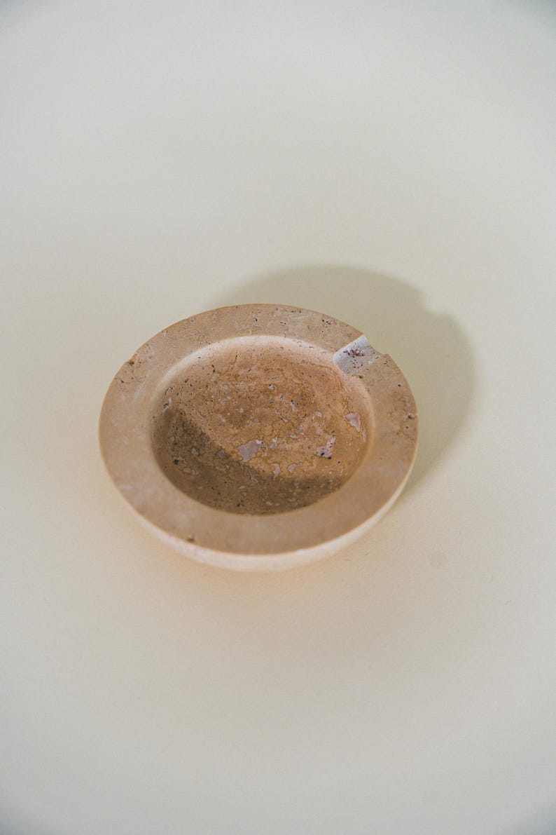 May include: A round, beige ashtray with a shallow bowl and a raised rim. The ashtray has a natural, textured appearance with a small notch for resting cigarettes. The ashtray is made of a light-colored material.