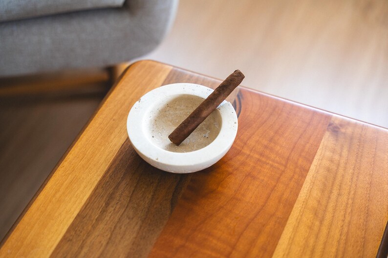 May include: A brown cigar rests in a small, off-white ashtray. The ashtray is circular and sits on a wooden table with a warm, reddish-brown finish. The scene suggests a moment of relaxation.