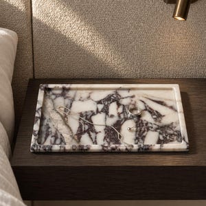 May include: A rectangular marble tray featuring a white base with prominent dark purple and grey veining. The tray holds a thin silver chain necklace, a simple silver band ring, and a pair of small silver stud earrings resting on its surface.