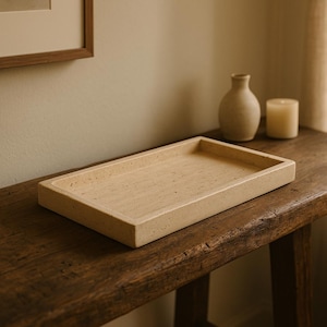 Puede incluir: Una bandeja rectangular de color beige claro hecha de piedra natural se encuentra sobre una mesa de madera rústica. La bandeja tiene una textura ligeramente rugosa y es perfecta para exhibir artículos decorativos.