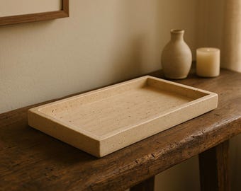 Puede incluir: Una bandeja rectangular de color beige claro hecha de piedra natural se encuentra sobre una mesa de madera rústica. La bandeja tiene una textura ligeramente rugosa y es perfecta para exhibir artículos decorativos.