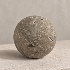 May include: A spherical stone object with a textured, mottled surface in shades of gray and brown. The surface features natural variations, small crevices, and swirling patterns throughout the rock, creating a rugged and organic appearance.