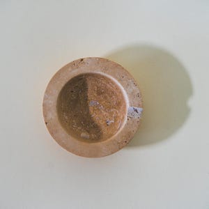 May include: A small, round, beige stone mortar with a circular bowl and a raised rim. The mortar is set against a light background, with a shadow cast to the side. A small sticker is visible on the rim.