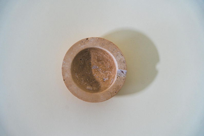 May include: A small, round, beige stone mortar with a circular bowl and a raised rim. The mortar is set against a light background, with a shadow cast to the side. A small sticker is visible on the rim.