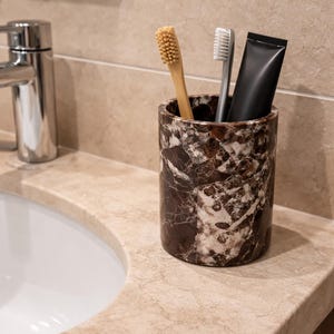 May include: A marble-patterned toothbrush holder with a bamboo toothbrush, a gray toothbrush, and a black tube of toothpaste. The holder sits on a beige countertop next to a white sink and a chrome faucet. The bathroom setting has a neutral color palette.