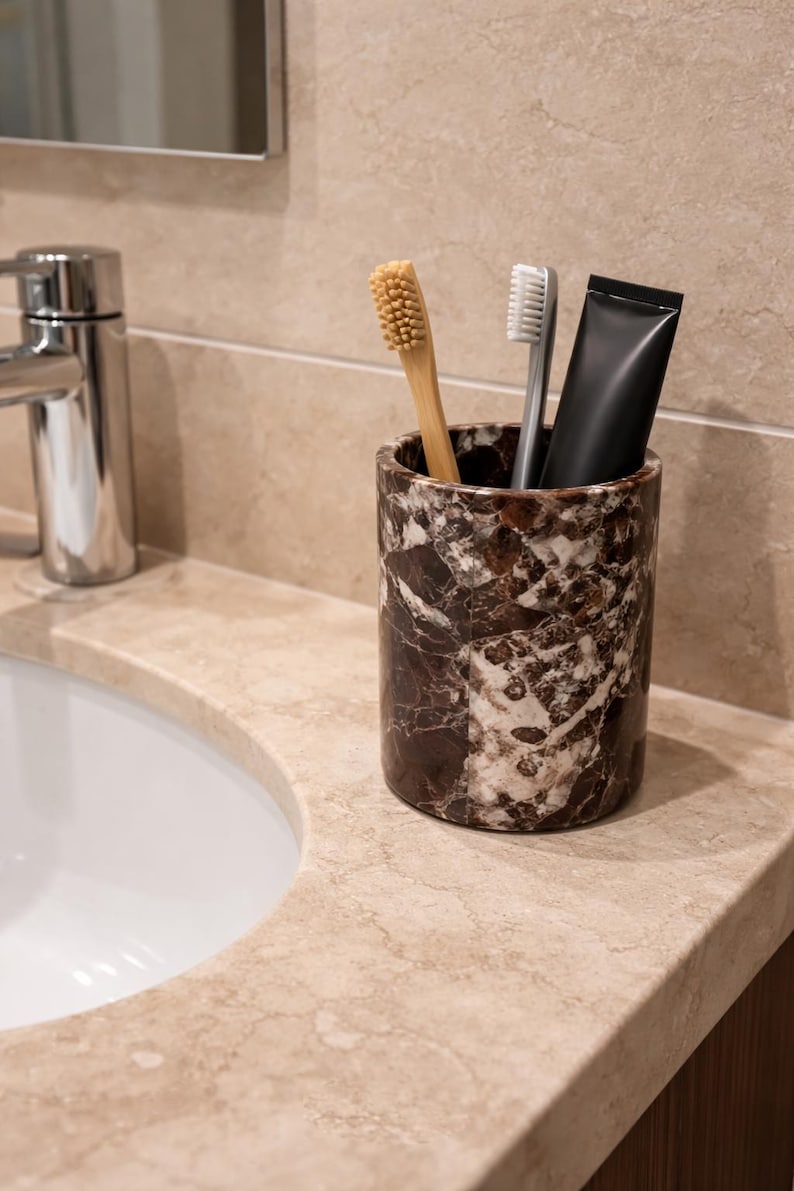 May include: A marble-patterned toothbrush holder with a bamboo toothbrush, a gray toothbrush, and a black tube of toothpaste. The holder sits on a beige countertop next to a white sink and a chrome faucet. The bathroom setting has a neutral color palette.