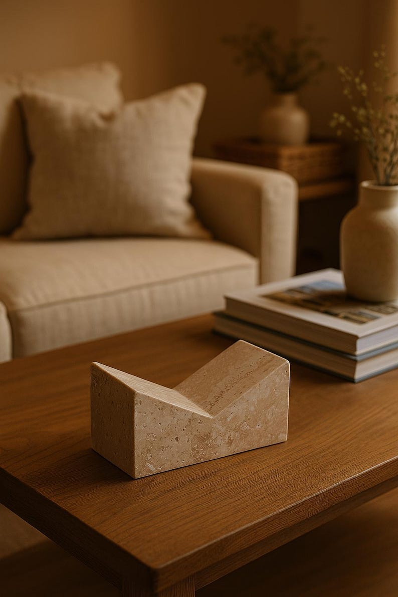 May include: A beige stone bookend with a V-shaped design sits on a wooden coffee table. The bookend is made of a textured stone material. A beige sofa and decorative vases are in the background.