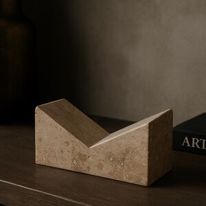 May include: A beige marble bookend with a V-shaped design. The bookend is made of a light-colored stone with a textured surface. The object is on a dark brown wooden surface.