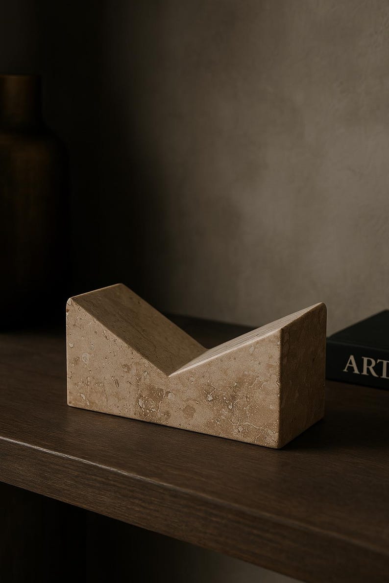 May include: A beige marble bookend with a V-shaped design. The bookend is made of a light-colored stone with a textured surface. The object is on a dark brown wooden surface.