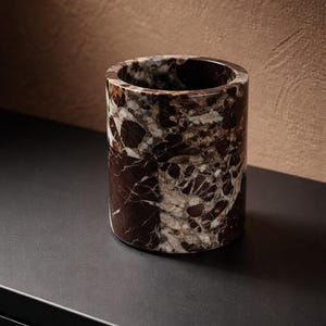 May include: A cylindrical container made of brown and white marble. The vessel has a smooth, polished exterior and a hollow interior. The marble pattern features dark brown veining against a lighter background. The container is likely used for holding items.