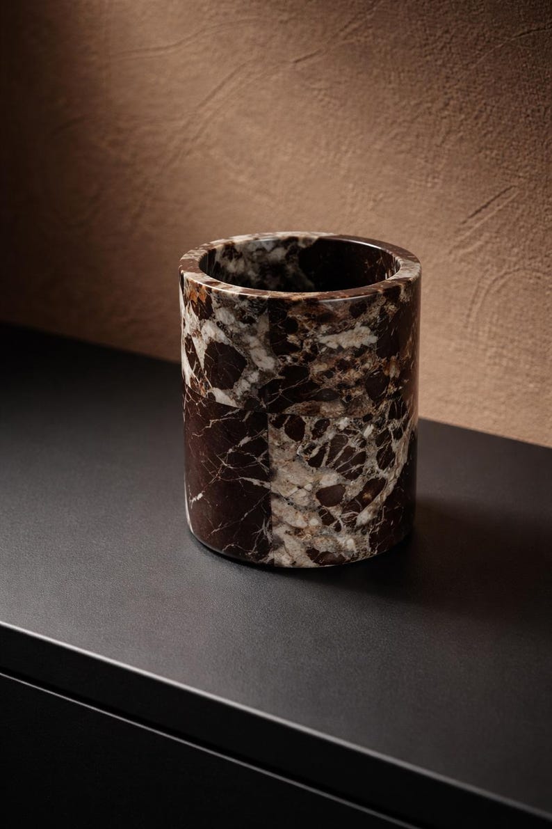 May include: A cylindrical container made of brown and white marble. The vessel has a smooth, polished exterior and a hollow interior. The marble pattern features dark brown veining against a lighter background. The container is likely used for holding items.