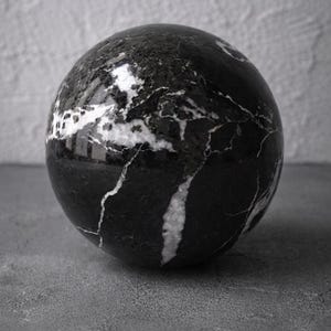 May include: A polished, spherical object with a black and white marble pattern. The orb is resting on a gray surface, with the light reflecting off its glossy finish. The veins of white marble create a striking contrast against the black.