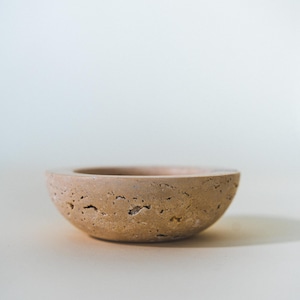 May include: A small, round, beige bowl with a textured surface. The bowl has a shallow depth and a wide rim. The neutral color and simple design make it suitable for various uses, such as a decorative piece or a small serving dish.
